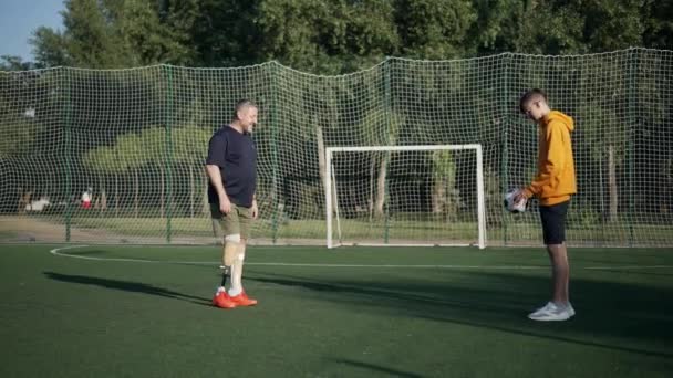 Široký záběr pozitivní zralé amputace a dospívající chlapec hrát fotbal ve zpomaleném filmu o slunném letním dni venku. Šťastné motivované Kavkazský otec a syn těší aktivní volný čas na slunci. — Stock video