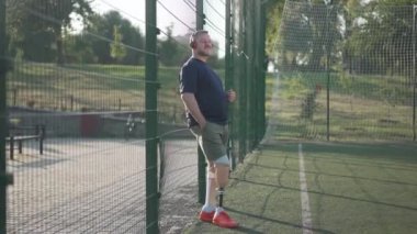 Geniş açılı portre mutlu beyaz erkek ampütasyon kulaklıkla müzik dinliyor yaz sporları sahasında çitlerin orada duruyor. Eğitimden sonra hobisinin keyfini çıkaran, protezli, olgun, kır saçlı bir adam..