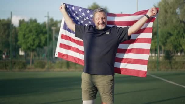 Inspirerad glad amputerad med amerikansk flagga tittar på kameran leende stående på sommar idrottsplan utomhus. Porträtt av leende idrottsman poserar på solig dag glädje jämställdhet. Långsamma rörelser. — Stockvideo