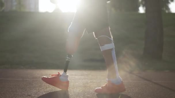 Visão lateral pernas protéticas de amputado masculino andando em raios solares em câmera lenta em pista de esportes vermelhos ao ar livre. Desportista caucasiano irreconhecível a passear ao sol. Conceito de motivação. — Vídeo de Stock