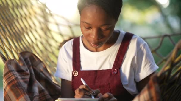 Сосредоточенная умная молодая афроамериканская женщина пишет ручкой, сидя на гамаке на улице. Пьедестал снял умный уверенный в себе студент-миллениал, обучающийся на солнечном свете. — стоковое видео