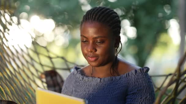 Χαμογελώντας όμορφη Αφροαμερικανή γυναίκα κάθεται στον ήλιο έξω με το βιβλίο εργασίας. Πορτρέτο της ενήλικης φοιτήτριας σπουδάζει την καλοκαιρινή μέρα κάθεται σε αιώρα. Νοημοσύνη και τρόπος ζωής. — Αρχείο Βίντεο