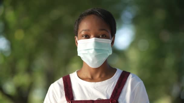 Portrét sebevědomé Afroameričanky v Covid-19 s maskou zkříženou na prsou, která v letním parku nestojí. Přední pohled štíhlá mladá dáma pózující venku na coronavirus pandemické ohnisko. — Stock video