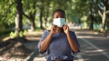 Covid-19 yüz maskeli Afro-Amerikalı genç bir kadın yaz parkında duran ciddi yüz ifadesine sahip kameraya bakıyor. Gün ışığında yavaş çekimde poz veren kendine güvenen bir kadının portresi.