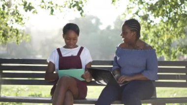 Yaz parkında dizüstü bilgisayar ve tabletle oturmuş ağır çekimde gülümseyen iki Afro-Amerikan öğrenci. Açık havada okuyan kendine güvenen güzel grup arkadaşlarının portresi..