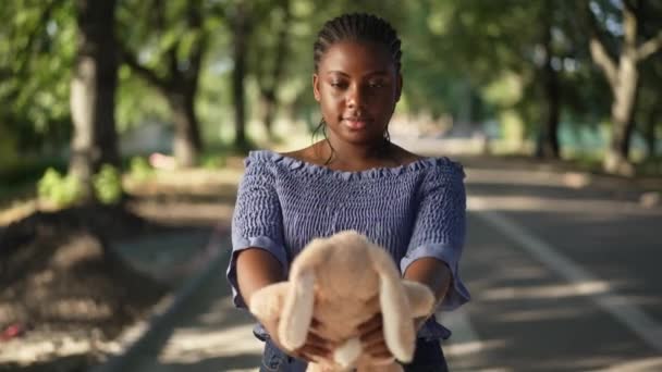 Счастливая афроамериканка тысячелетия любуется игрушечным кроликом, стоящим в солнечном парке на открытом воздухе. Портрет положительной красивой женщины, наслаждающейся летними выходными в замедленной съемке. — стоковое видео