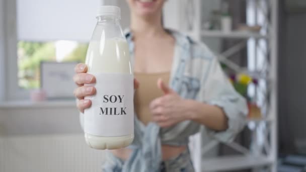 Yakın plan soya sütü şişesi ve arka planda gülümseyen beyaz bir kadın. Genç, sıska ve mutlu bayan evde sağlıklı süt içecekleriyle poz veriyor.. — Stok video