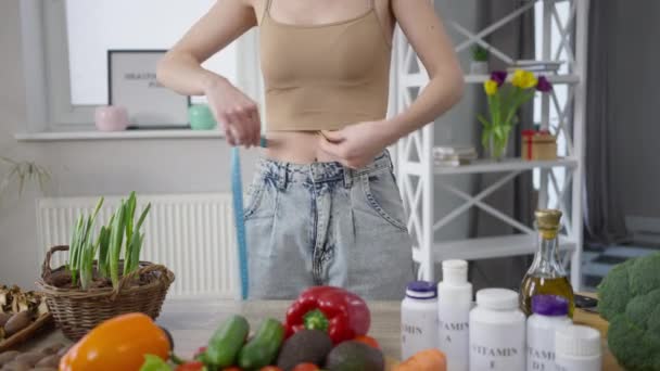 Unrecognizable slim woman measuring waist standing at table with healthful vegan food indoors. Young slender Caucasian millennial lady control calories and body weight. Healthy lifestyle. — Stock Video