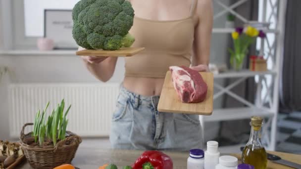 Front view unrecognizable woman choosing broccoli or beef steak for dinner. Young slim Caucasian lady standing at table holding healthy and unhealthy food thinking. Choice concept. — Stock Video