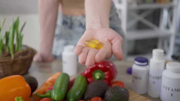 Крупным планом женский кулак открывает ладонь с желтыми витаминами за столом со здоровыми овощами. Неузнаваемая юная стройная белая женщина показывает диетическую добавку в помещении дома. — стоковое видео