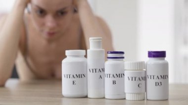 Arka planda bulanık beyaz kadın düşüncesi olan bir dizi vitamin şişesi masanın üzerinde. İçinde hapları seçen bin yıllık ince, güzel bir bayanın bulunduğu kapalı alanlarda ilaç takviyesi..