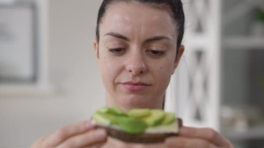 Sıska vejetaryen bir kadının portresi sağlıklı çavdar ekmekli sandviçi ısırıyor ve salatalık gülümsüyor. Yakın plan mutlu Kafkasyalı vejetaryen kadın evde sağlıklı yiyecekler yiyor ve tadını çıkarıyor..