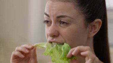 Yeşil salata yaprağı yiyen ve tadını çıkaran mutlu vejetaryen kadının yakın plan yüzü. Sağlıklı yiyecekler çiğneyen, gülümseyen binlerce yıllık beyaz kadının portresi..