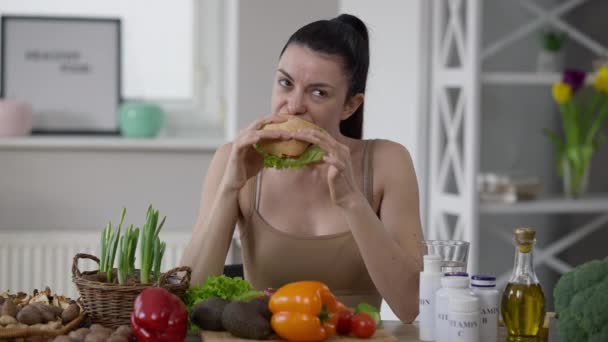 Střední střela mladá štíhlá běloška veganská ochutnávka lahodného nezdravého hamburgeru se spokojeným výrazem ve tváři. Portrét krásné dámy jíst nezdravé jídlo sedí u stolu se zeleninou. — Stock video