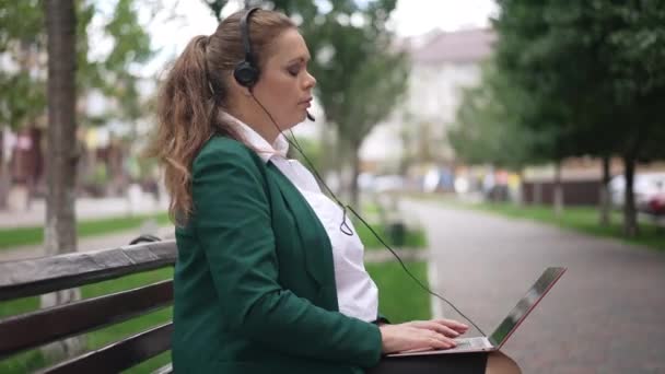 Vista lateral con confianza exitosa mujer de negocios saludando en el chat de vídeo portátil hablando sentado en el banco de la ciudad. Retrato del experto consultor comercial caucásico de tamaño grande en mensajería de auriculares en línea. — Vídeos de Stock