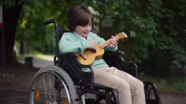 Yetenekli, tekerlekli sandalyedeki engelli, açık havada, yaz parkında ukulele çalan beyaz çocuk. Motive olmuş mutlu bir çocuğun tek başına hobisinin portresi. Yalnızlık ve sakatlık kavramı.