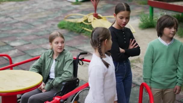 Skupina přátel mluví ignorovat postiženou smutnou dívku na vozíčku na hřišti. Kavkazský chlapec a dívky si nevšímají rozrušených nemocných dětí na veselé procházce. Koncept ignorace a hrubosti. — Stock video