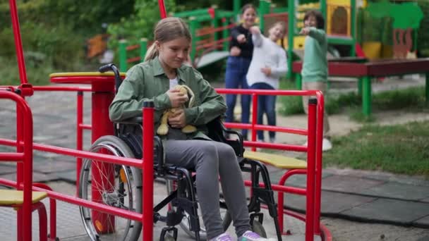 Плачущая отчаявшаяся белая девушка в инвалидной коляске обнимает игрушку, а размытые дети смеются над фоном. Портрет запуганного ребенка-инвалида на детской площадке с грубыми детьми. Медленное движение. — стоковое видео
