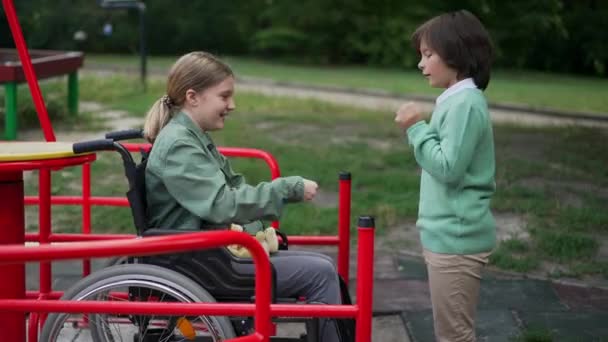 Vue de côté joyeux garçon et fille caucasien en fauteuil roulant jouer ciseaux en papier de roche à l'extérieur sur aire de jeux. Amusez-vous bien chez des amis joyeux et joyeux. Mode de vie et handicap des enfants. — Video