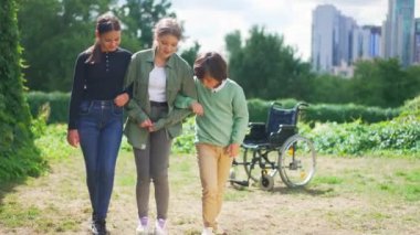 Geniş açılı arkadaşlar, arka planda tekerlekli sandalyeyle gün ışığında yürümeye çalışan hasta kıza yardım ediyorlar. Gülümseyen Kafkasyalı erkek ve kız arkadaşa destek veriyorlar..