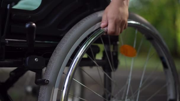 Close-up child hand rolling wheel of wheelchair in slow motion leaving. Unrecognizable Caucasian little disabled boy riding manual assistive device outdoors in summer park. — Stock Video
