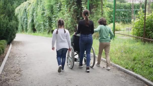 Kafkasyalı çocuklar tekerlekli sandalye itiyor ve engelli arkadaşları dışarıda ağır çekimde yürüyorlar. Geniş açılı mutlu çocuk ve kızlar felçli kızla ilgileniyorlar yaz parkında geziniyorlar. — Stok video