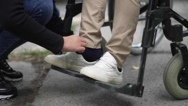 Cuidado irreconocible Niña adolescente caucásica poniéndose zapatos en los pies de un amigo discapacitado en silla de ruedas. Amable hermana cuidando del parapléjico hermano paralítico al aire libre. Movimiento lento. — Vídeos de Stock