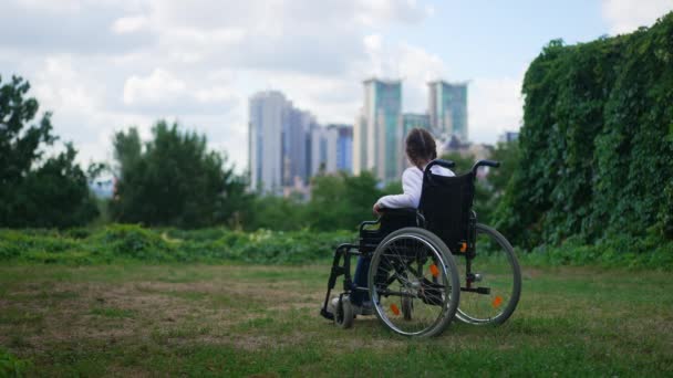 Szeroki letni park z niepełnosprawną białą dziewczyną na wózku inwalidzkim podziwiającą piękno miejskiego miasta w tle. Widok z tyłu sparaliżowane dziecko samotnie na zewnątrz patrząc daleko myśląc. Zwolniony ruch. — Wideo stockowe