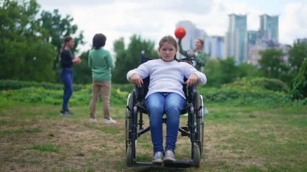 Brede shot depressief gehandicapt meisje in rolstoel kijken naar camera met droevige gezichtsuitdrukking als vrolijke kinderen spelen bal op de achtergrond. Portret van gefrustreerd wanhopig kind buiten in het park. — Stockvideo