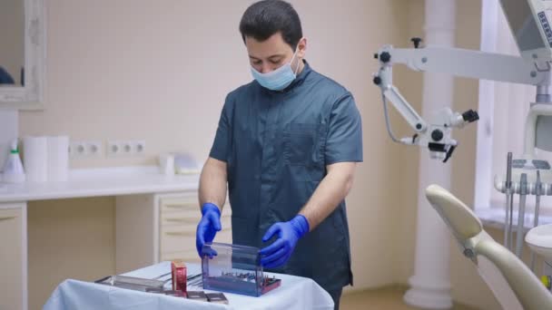 Dentista do Oriente Médio masculino abrindo kit cirúrgico dentário em câmera lenta em pé no escritório dentro de casa. Especialista concentrado em máscara facial a trabalhar no hospital. Cirurgia oral e medicina conceito. — Vídeo de Stock