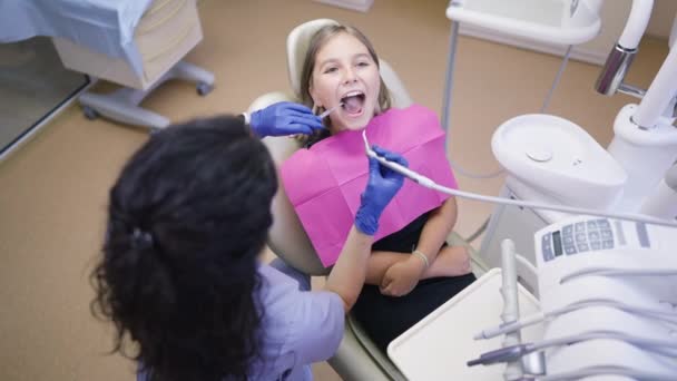 Vysoký úhel pohledu dospívající dívka v zubař židle otevření úst jako lékař pomocí ústní zrcadlo a vysoušeč zubů ve zpomaleném filmu. Profesionální běloška vyšetřující zuby pacienta na klinice. — Stock video