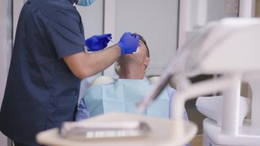 Profesyonel ortodontist olarak dudak retraktörü ile dişçi koltuğundaki yetişkin erkek hasta aletleri kullanarak ağız boşluğunu inceliyor. Hastane kliniğinde kendinden emin beyaz bir adam ve Orta Doğulu bir doktor var..
