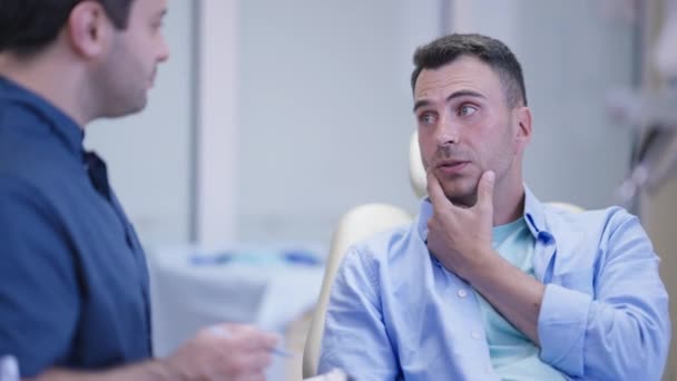 Portrait of handsome Caucasian man talking with Middle Eastern doctor sitting in dentist chair in hospital. Worried male patient complaining toothache indoors in slow motion. Orthodontist visit. — Stock Video