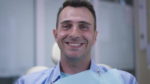 Передня сторона виглядає задоволеною кавказькою брюнеткою з блакитними очима і зубатою посмішкою, дивлячись на камеру, що посміхається. Портрет із зображенням веселих чоловіків у стоматологічному офісі, що вихваляють здорові зуби.. — стокове відео