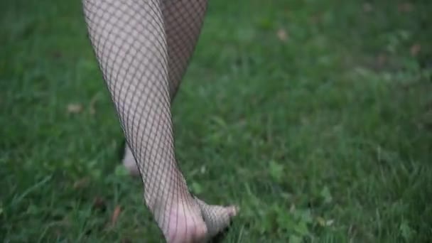 Tracking schot barefoot slanke jonge dronken vrouw in visnet kousen lopen op groen gras passeren lege alcohol fles verspreid hoge hakken en portemonnee. Dronken blanke dame wandelen op het gazon in het park. — Stockvideo