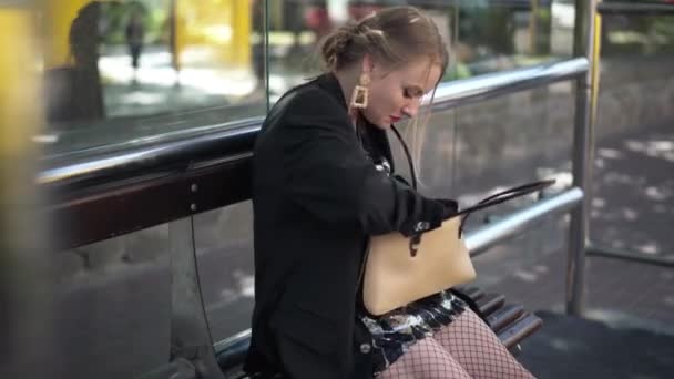 Junge schöne Frau auf der Suche nach Coronavirus-Gesichtsmaske sitzt an der Bushaltestelle und geht weg. Zuverlässige kaukasische Dame, die nach Ausbruch der Covid-19-Pandemie zum Transport in die Stadt aufbricht. — Stockvideo