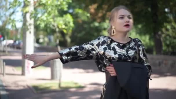 Joven elegante mujer delgada autoestop en la carretera urbana al aire libre por la mañana. Retrato de la hermosa dama caucásica en el soleado día de verano en el parque solicitando un paseo. Concepto de estilo de vida. — Vídeos de Stock