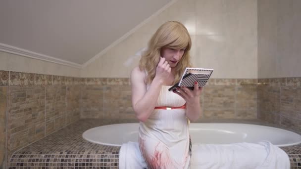 Blond Caucasian LGBTQ trans person applying face powder looking at camera smiling. Portrait of confident positive transgender woman in elegant dress sitting on bathtub at home posing indoors. — 图库视频影像