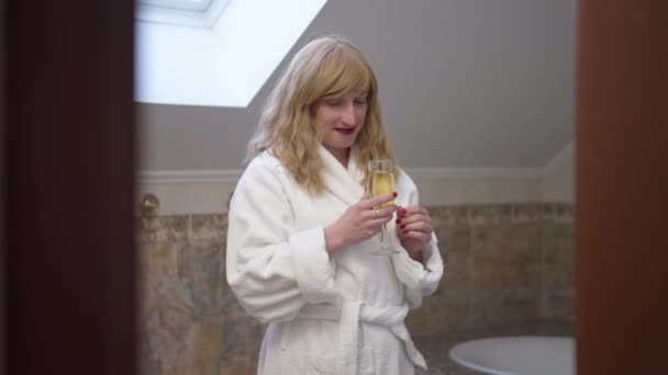 Розслаблена безтурботна трансгендерна жінка в халаті насолоджується смаком білого вина, що стоїть у ванній кімнаті. Портрет щасливої упевненості Кавказької дивної людини, яка п'є алкоголь вдома. — стокове відео