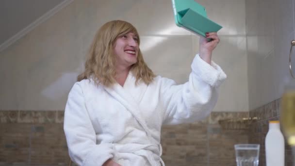 Uśmiechnięta, szczęśliwa transwestyta machająca na tablecie, siedząca w łazience w domu. Portret pewnej siebie osoby kaukaskiej LGBTQ blogującej w domu na czacie online w mediach społecznościowych za pomocą czatu wideo. — Wideo stockowe