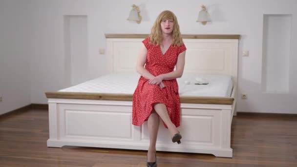 Foto ampla retrato confiante mulher LGBT trans em vestido vermelho pontilhado e couro preto de salto alto sentado em cama confortável dentro de casa. Loira caucasiana transgênero pessoa no quarto luxuoso. — Vídeo de Stock