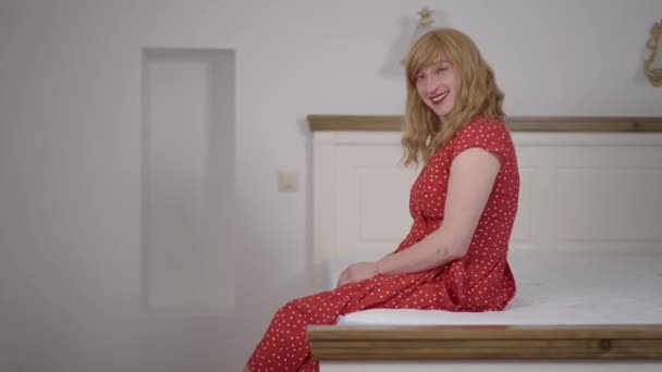 Seitenansicht lächelnd glückliche Transgender LGBTQ-Person auf dem Bett sitzend und in die Kamera blickend. Porträt einer fröhlichen, selbstbewussten kaukasischen Transfrau im rot gepunkteten Kleid, die drinnen im Schlafzimmer posiert. — Stockvideo