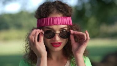 Kameraya bakarak güneş gözlüğü takan, gülümseyen beyaz kadın. Vesikalık portre, neşeli, kendine güvenen, saç bandı takmış çekici bir kadın güneşli yaz parkında poz veriyor..