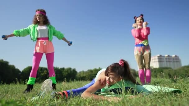 Slank brunette fleksibel sportskvinne som strekker muskler som venner i retroklær som trener i bakgrunnen. Selvsikre, hvite, vakre, unge kvinner som trener i solskinn på grønne sommerplener. – stockvideo