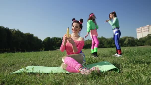 Weitschossene glückliche Frau beim Bananenschälen auf einer Matte sitzend, während verschwommene Frauen lachend zeigen und sich über Freundin lustig machen. Schlanke schöne kaukasische Retro-Lady beim Outdoor-Training mit scheinheiligen Freunden. — Stockvideo