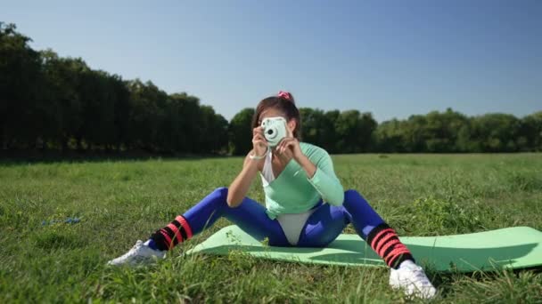Large plan heureuse femme rétro talentueuse prenant des photos sur l'appareil photo assis sur tapis d'exercice sur prairie verte d'été. Femme sportive caucasienne excitée photographe jouissant de passe-temps en plein air dans les années 1990. — Video