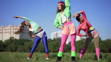 Yeşil çayır üzerinde güneş ışığında ısınan üç beyaz kadın eskimiş sporcunun portresi. Geniş açı, 1990 'larda güneşli bir günde egzersiz yapan kendinden emin, zayıf kadınlara yönelik. Yavaş çekim.