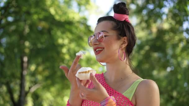 역으로 된 옷을 입은 젊은 코 켓 백인 여성 이 여름 공원에서 웃으며 서서 느린 동작으로 맛있는 도넛 크림을 맛보고 있다. 매력적 인 아름다운 여인 이 디저트를 먹으며 웃고 있다. — 비디오