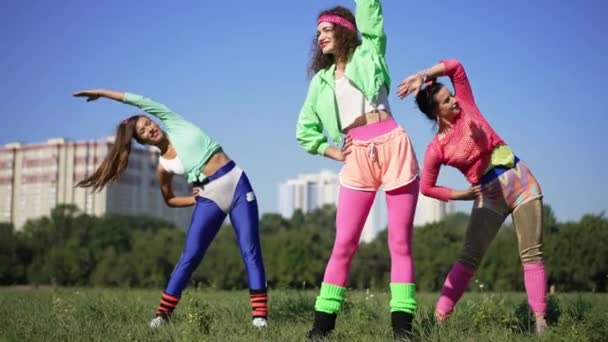 Portrét tří zdatných kavkazských retro sportovkyň ohýbajících se ve slunečním světle venku na zelené louce. Široký záběr sebevědomé štíhlé ženy cvičení v devadesátých letech za slunečného dne. Zpomalený pohyb. — Stock video