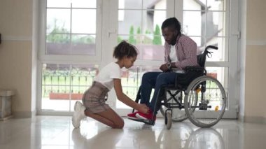 Geniş açı pozitif Afro-Amerikalı kız, engelli bir babaya bakıyor, ayakkabı bağlıyor. Yan görünüm, gülümseyen kız ve tekerlekli sandalyedeki adam evde ayakkabılarını giyip konuşuyor..